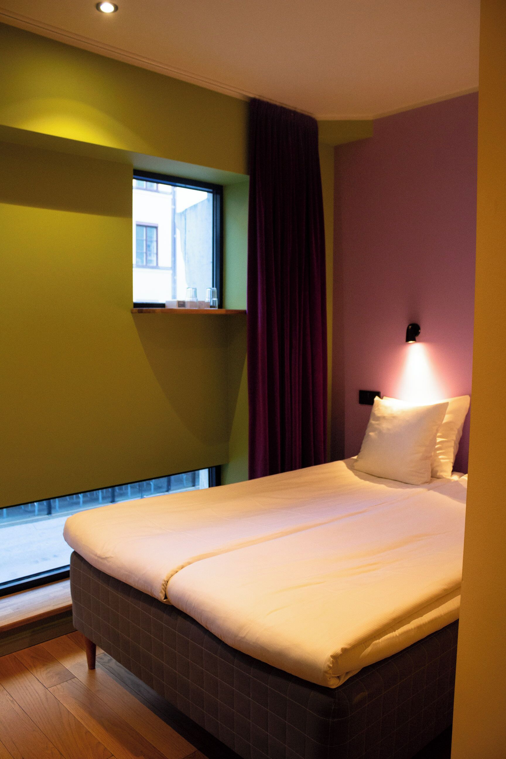 A hotel room with a bed, a window, and a wall-mounted light fixture.