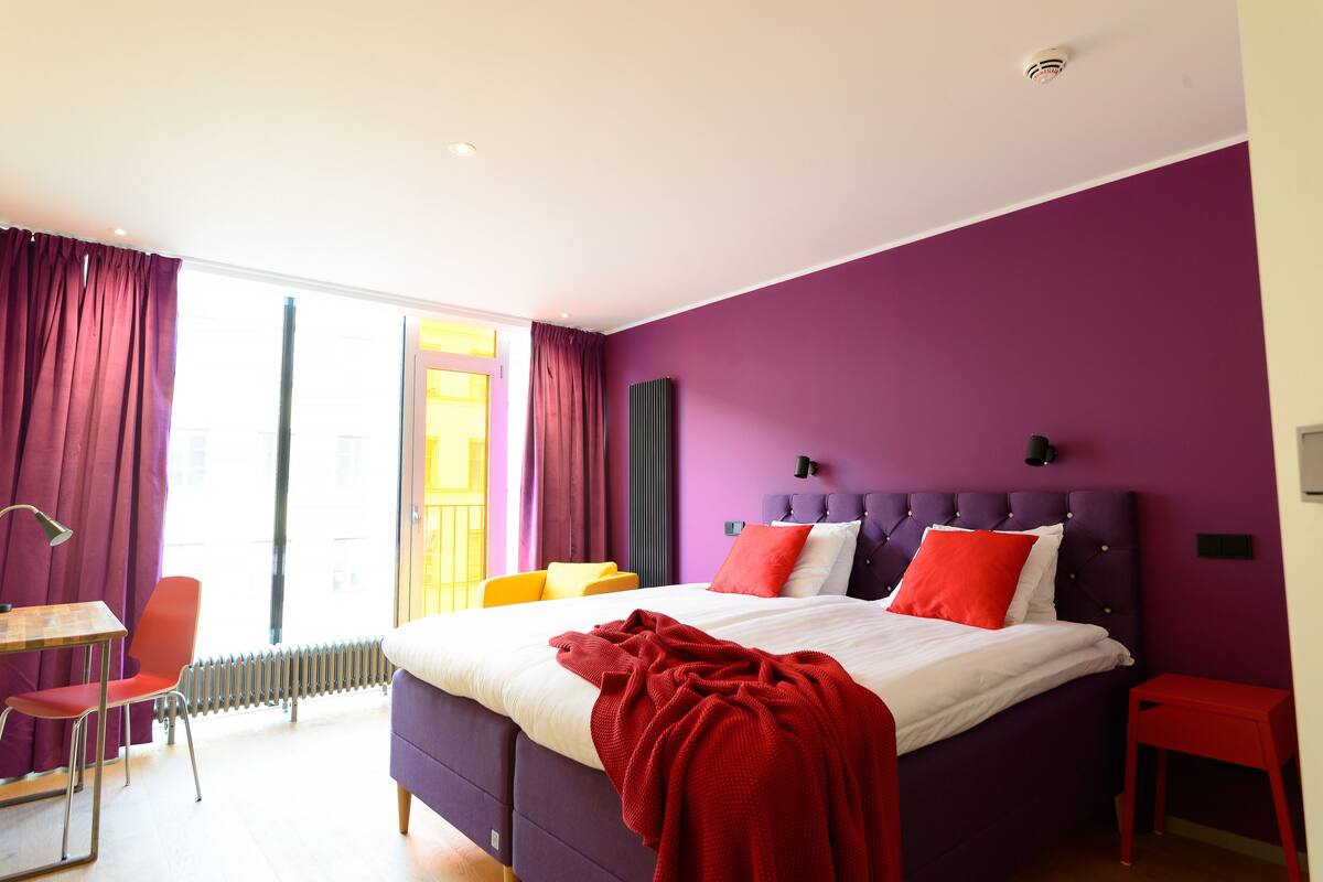 Una camera da letto moderna con una testiera viola, una coperta rossa e vista sulla città attraverso ampie finestre.