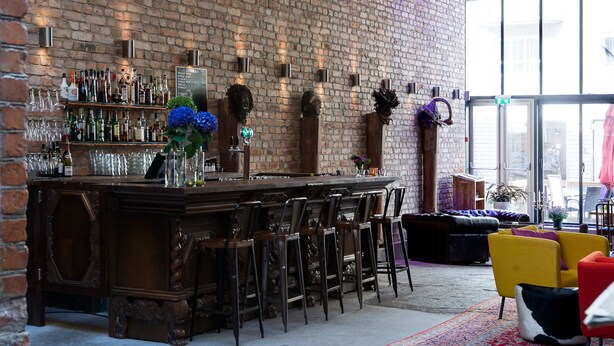 A bar area with a brick wall, wooden bar counter, and multiple stools. - Hellstens Glashus (Stockholm)