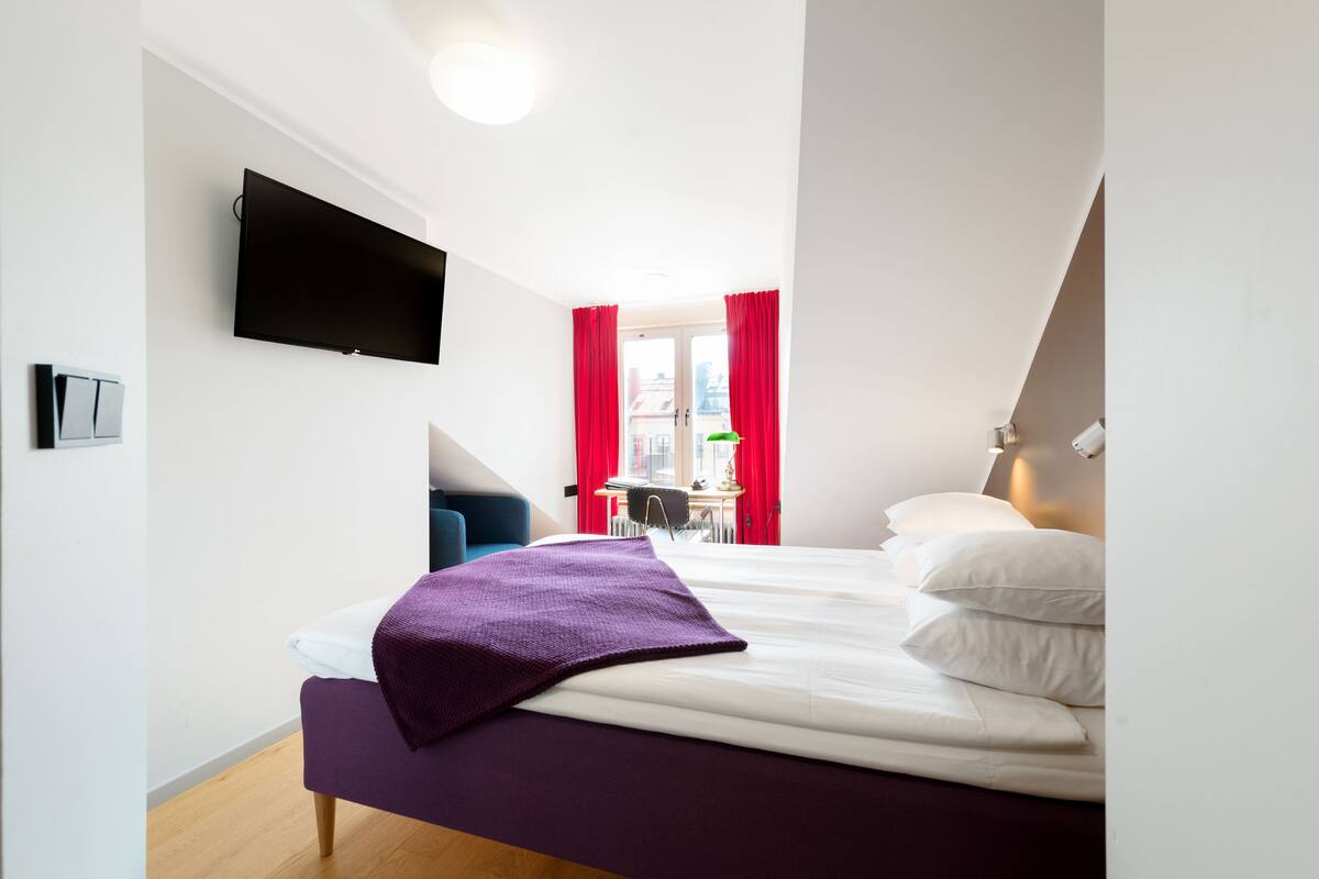 Una camera d'albergo moderna con un letto, una TV a schermo piatto fissata al muro e una scrivania con una sedia sullo sfondo.
