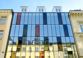 Un edificio moderno con amplios ventanales que reflejan el cielo y edificios vecinos.