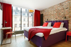 Un dormitorio moderno con cama púrpura, cortinas rojas y pared de ladrillo.