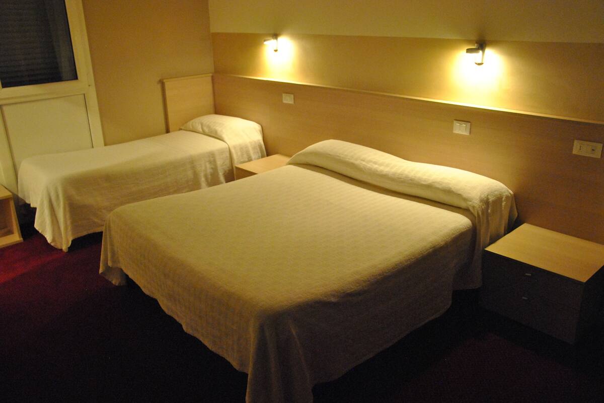 Une chambre d’hôtel avec deux lits simples et un grand lit, tous dotés de linge de lit blanc. La chambre comprend des tables de chevet et des lampes fixées au mur.