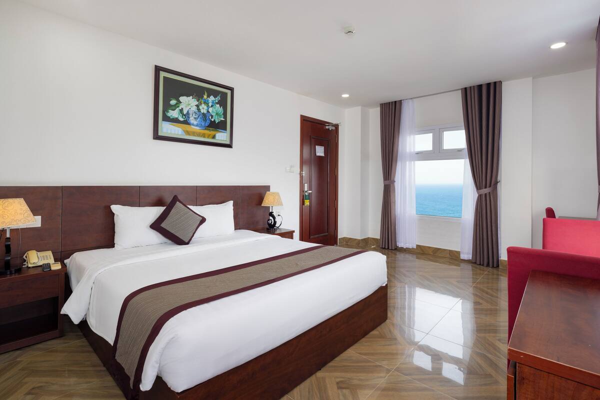 A hotel room with a large bed, wooden headboard, a painting on the wall, a desk with a chair, and a view of the ocean through the window.