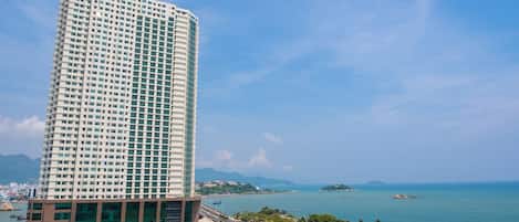 A tall, modern hotel with a glass facade, located near a beach with a clear view of the sea and distant mountains.