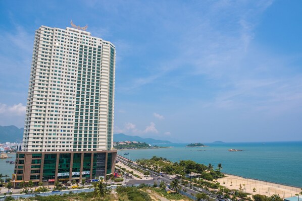 A tall, modern hotel with a glass facade, located near a beach with a clear view of the sea and distant mountains.