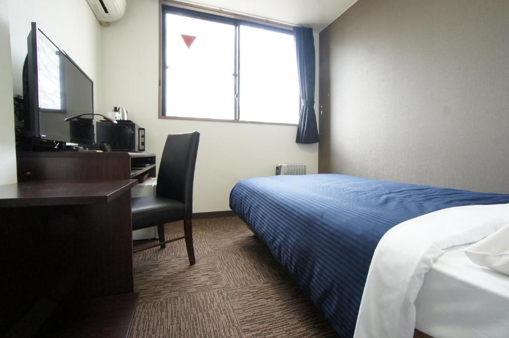 A hotel room with a bed, a desk with a computer, a chair, and a window with curtains.
