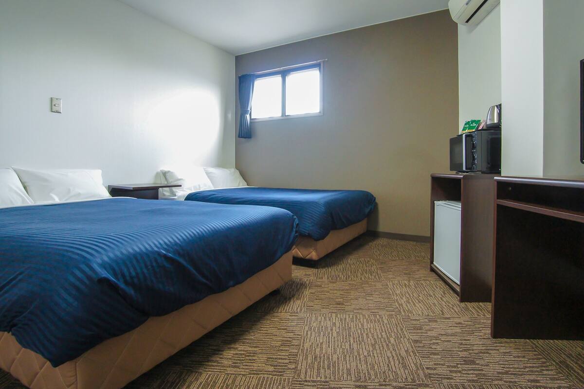 A hotel room with two beds, a window with curtains, a desk with a microwave, and an air conditioning unit.