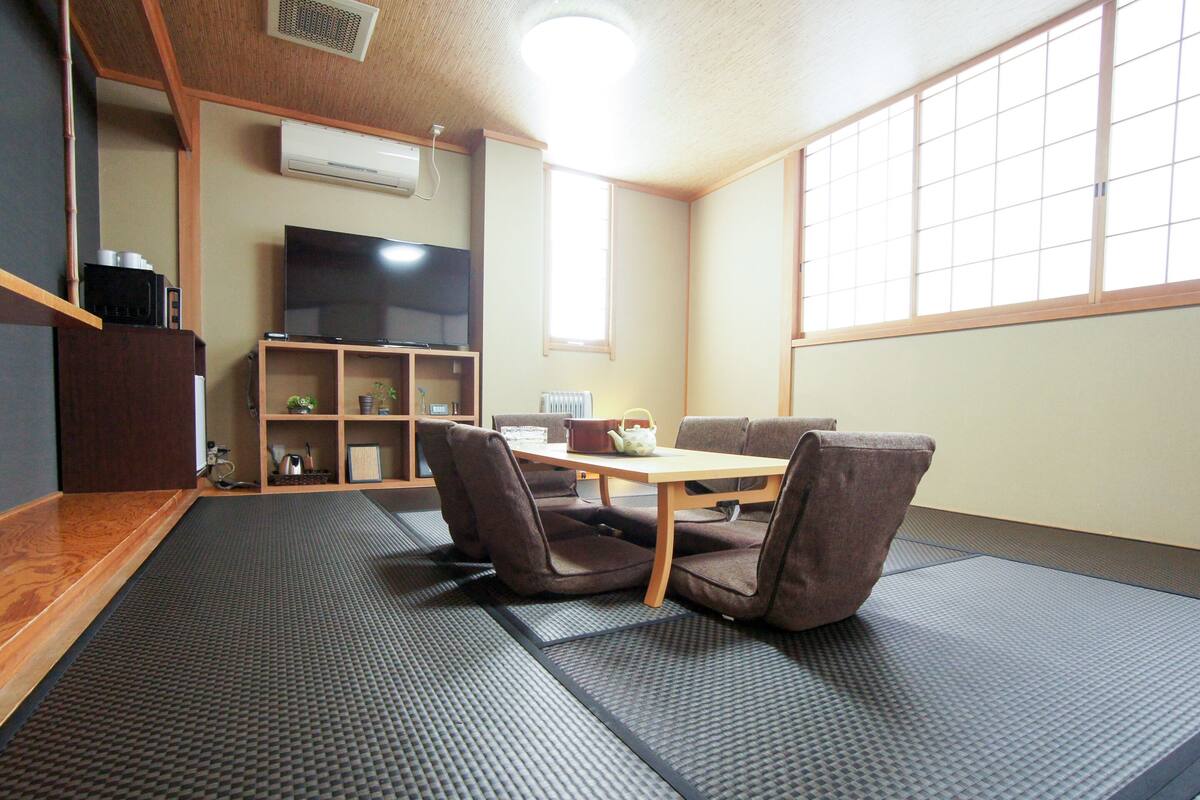 A room with tatami mats, a low table, and four chairs. There is a television on the wall, a shelf with items, and a window with a view.