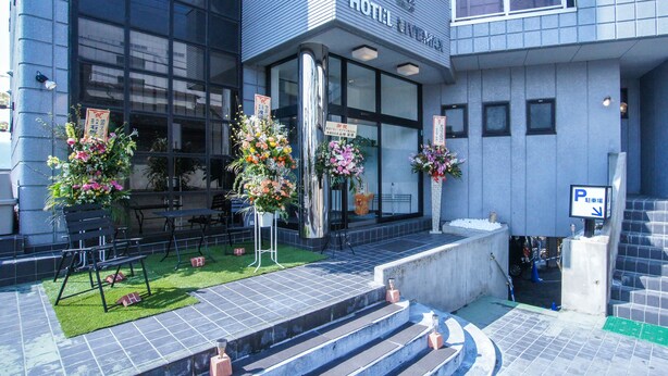 En hotelindgang med en moderne facade, trapper der fører op til indgangen, og blomsterarrangementer ved foden.