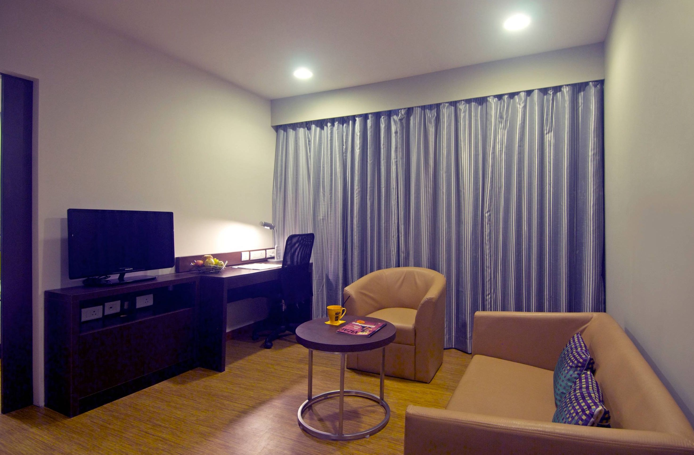 Habitación de hotel moderna con televisor de pantalla plana, escritorio con silla, una mesita redonda con una revista, sofá y un ventanal con cortinas.