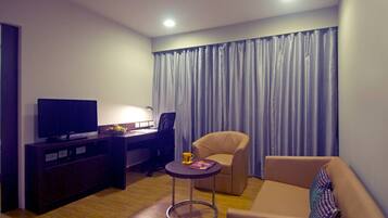 Habitación de hotel moderna con televisor de pantalla plana, escritorio con silla, una mesita redonda con una revista, sofá y un ventanal con cortinas.