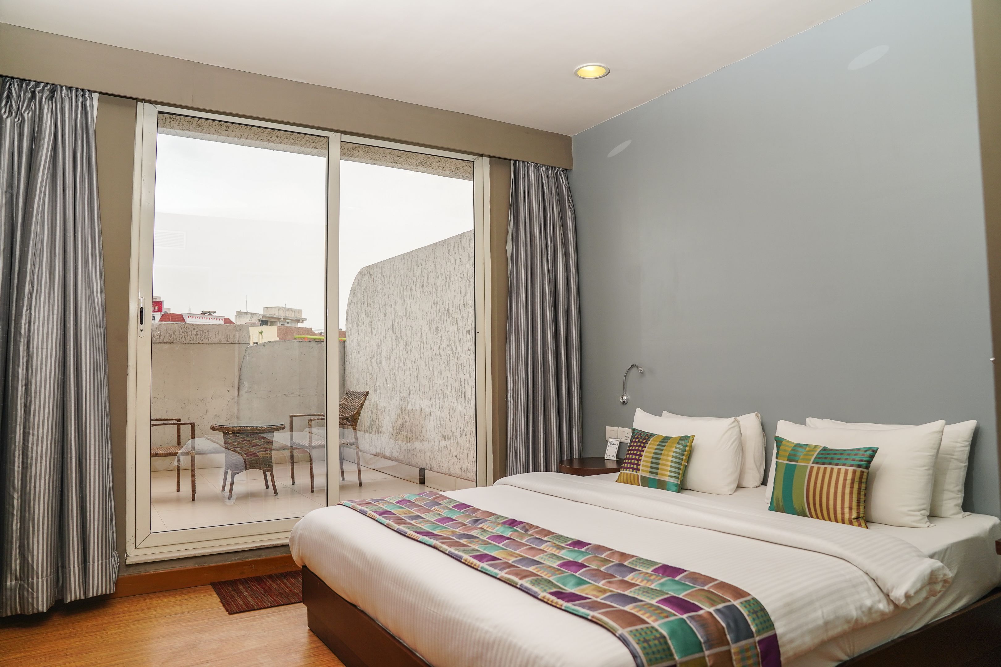 A hotel room with a large bed, a view of the city through a sliding door, and a balcony with a table and chairs.