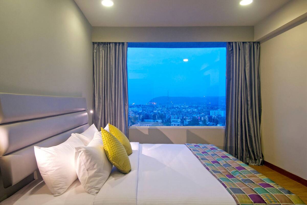 A hotel room with a large bed, a view of a cityscape through a window, and curtains.