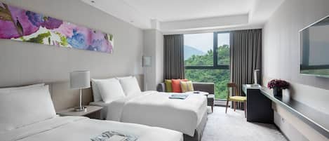 A hotel room with two beds, a desk, a chair, a TV, and a large window with a view of greenery.