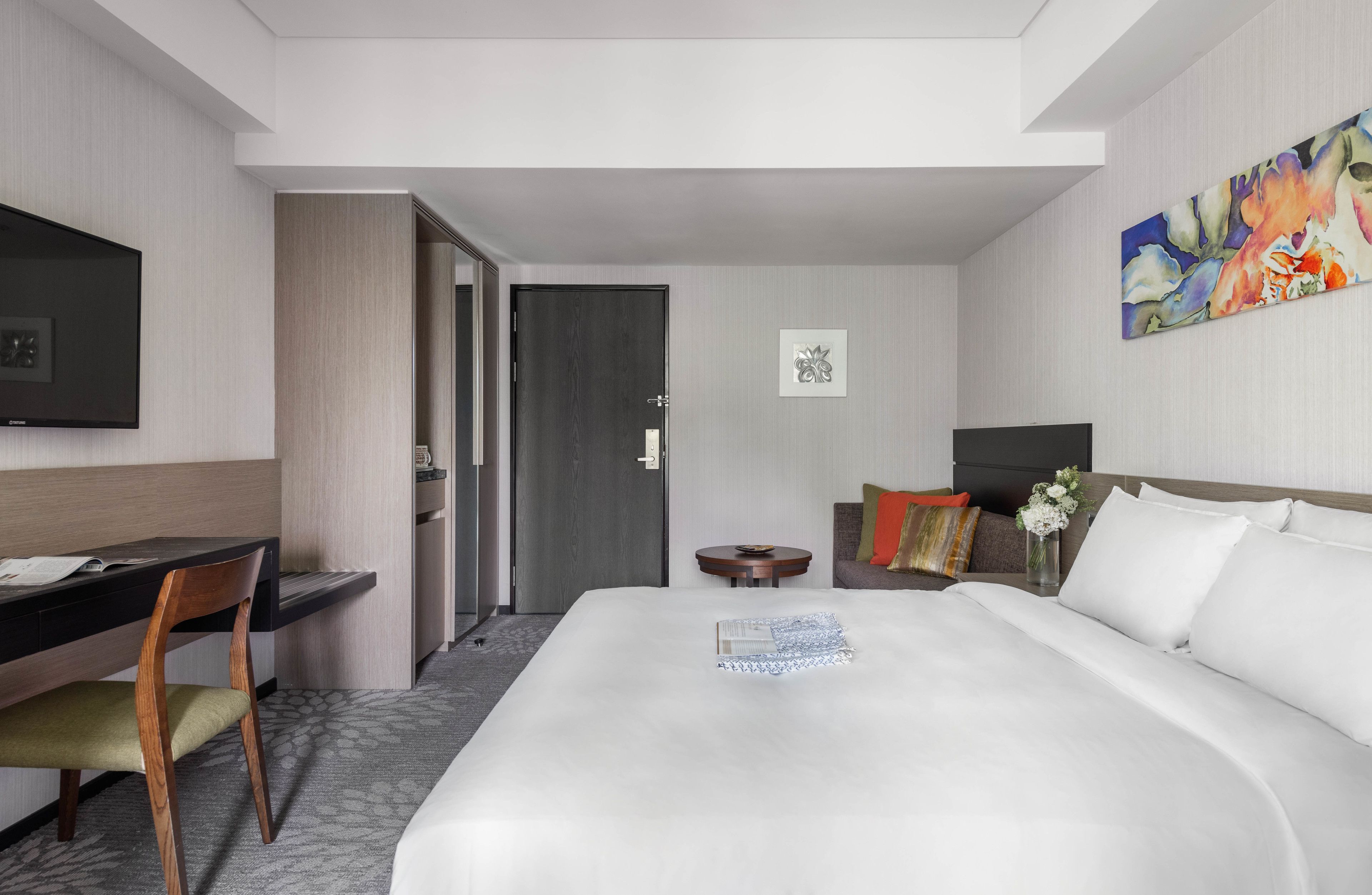 A hotel room with a bed, a desk, a chair, a TV, and a painting on the wall.