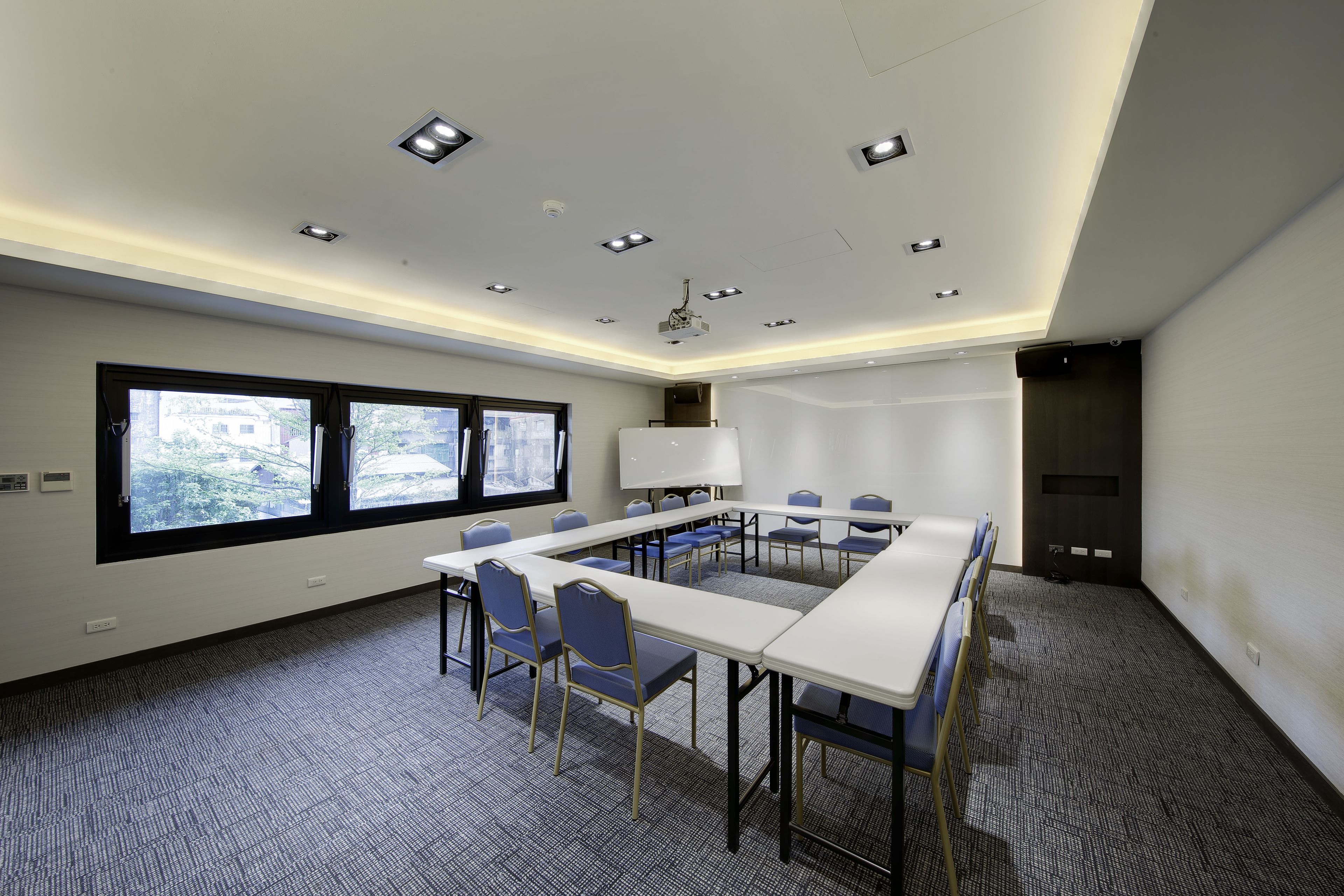 Un salón de conferencias con una larga mesa, sillas, una pantalla de proyector y una ventana con vistas a edificios.