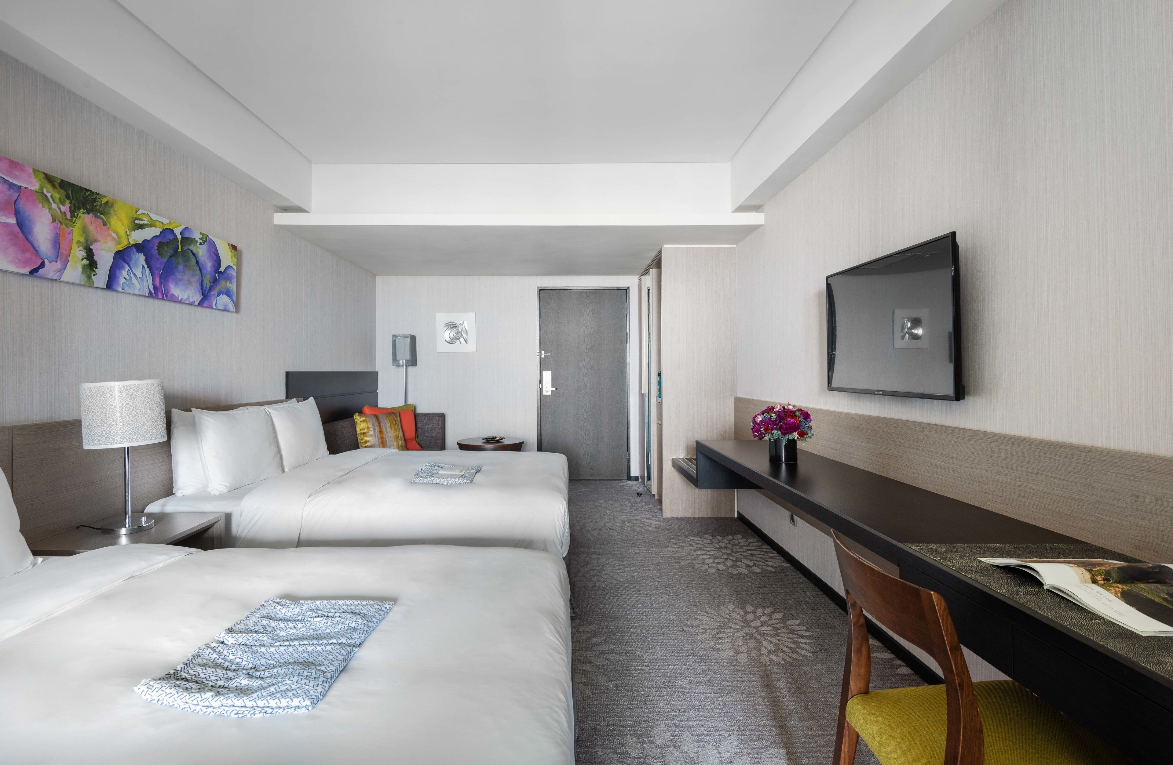 A hotel room with two beds, a desk, a chair, a TV, and a door leading to another room.