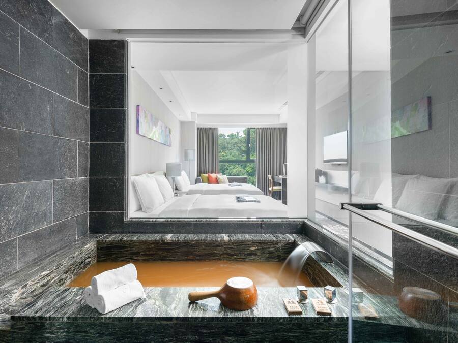 A modern bathroom with a large bathtub, a shower, and a glass-enclosed shower area. There is a bed with a view of greenery outside.