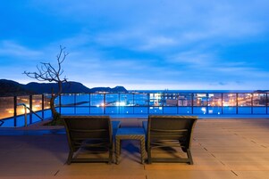 Una terraza con dos sillones de mimbre y una mesa, con vistas a un paisaje urbano que incluye una masa de agua y montañas al fondo.