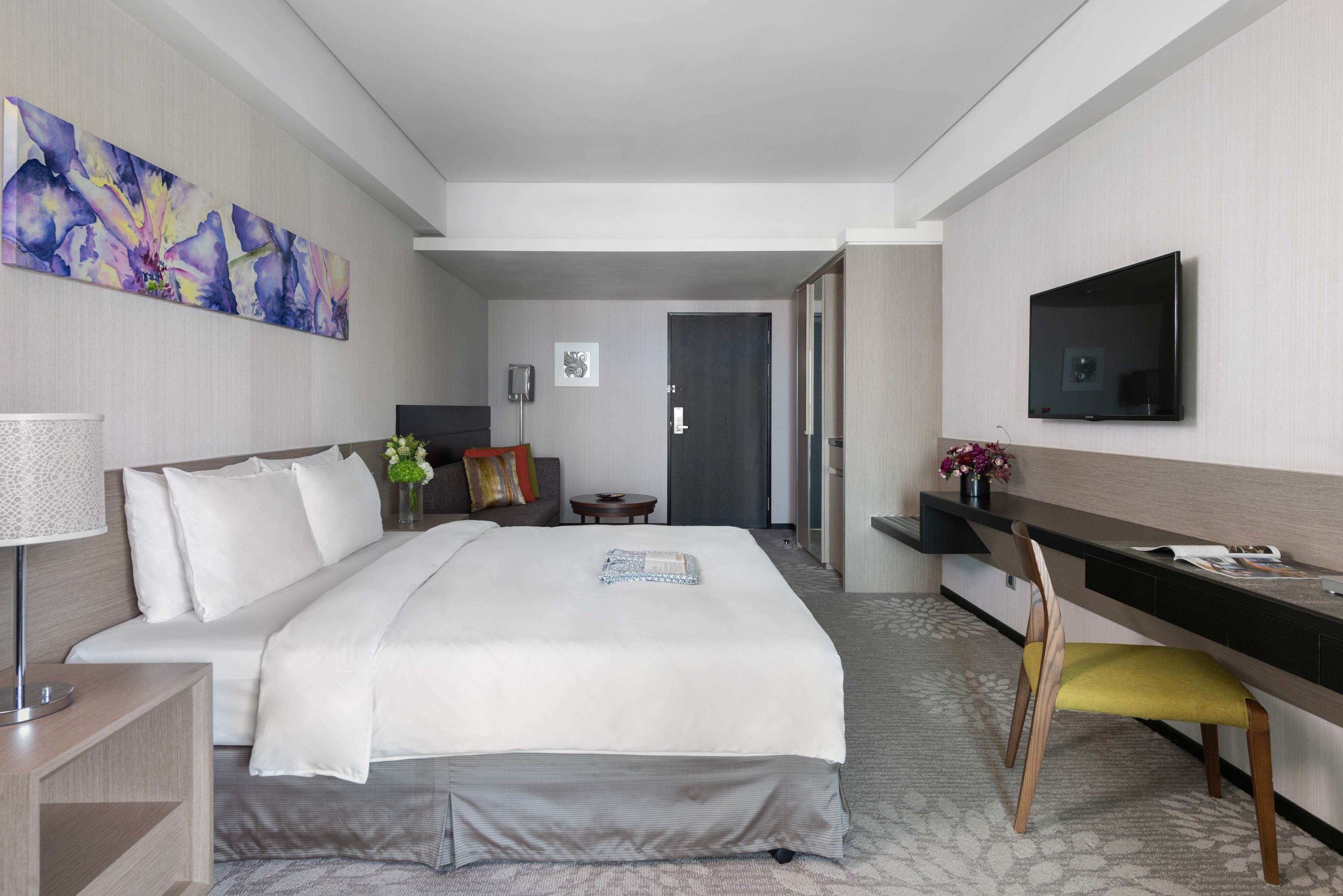 A hotel room with a bed, bedside table, lamp, TV, desk, chair, and artwork on the walls.