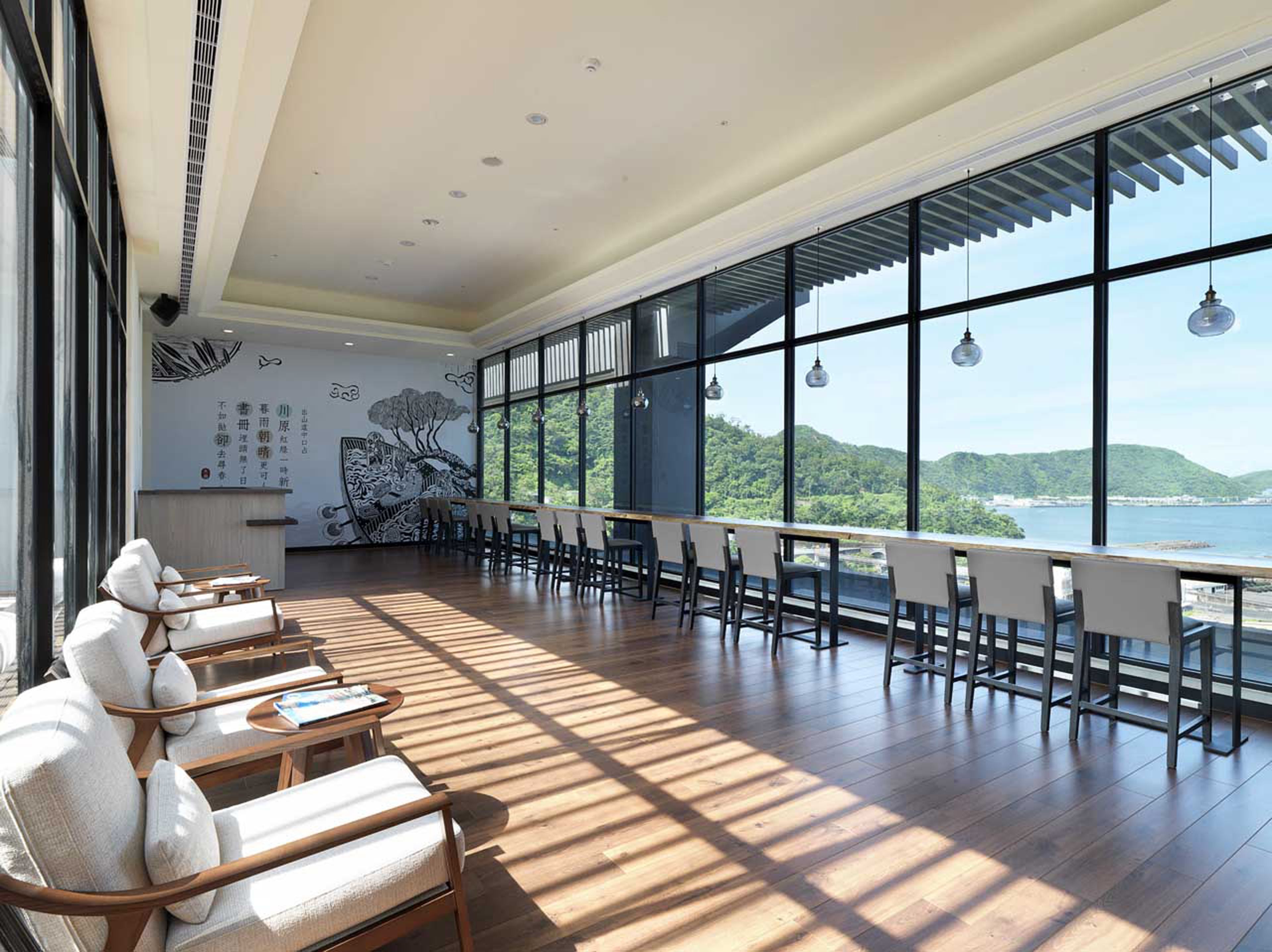 Un espacio interior amplio con una larga mesa de madera y sillas, ventanas grandes que ofrecen vistas a un paisaje costero y un mural en la pared.