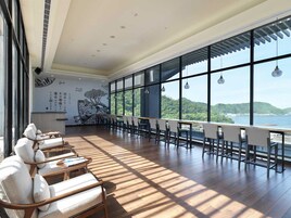 Un espacio interior amplio con una larga mesa de madera y sillas, ventanas grandes que ofrecen vistas a un paisaje costero y un mural en la pared.