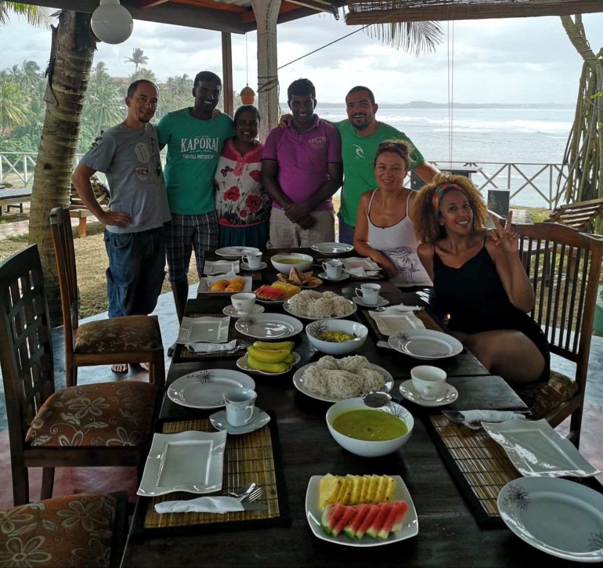A group of people seated at a table with a variety of dishes, including rice, vegetables, and fruit, set up outdoors with a view of the ocean.