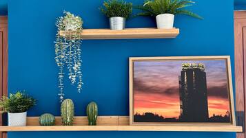 Un mur bleu orné d’une étagère en bois sur laquelle sont disposées différentes plantes, ainsi qu’un tableau représentant un bâtiment au coucher du soleil.