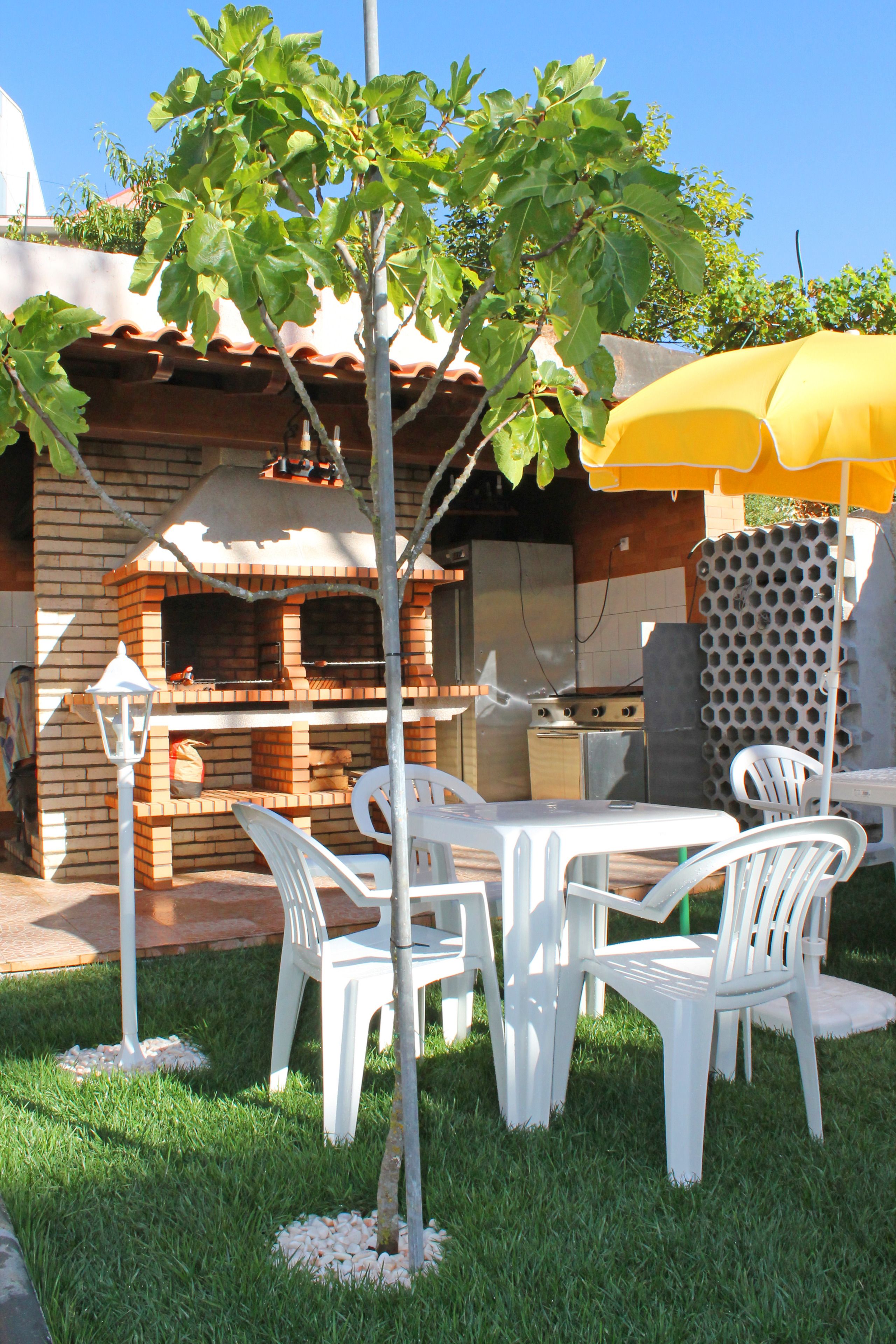 A brick barbecue grill with a chimney, surrounded by white plastic chairs and a yellow umbrella.