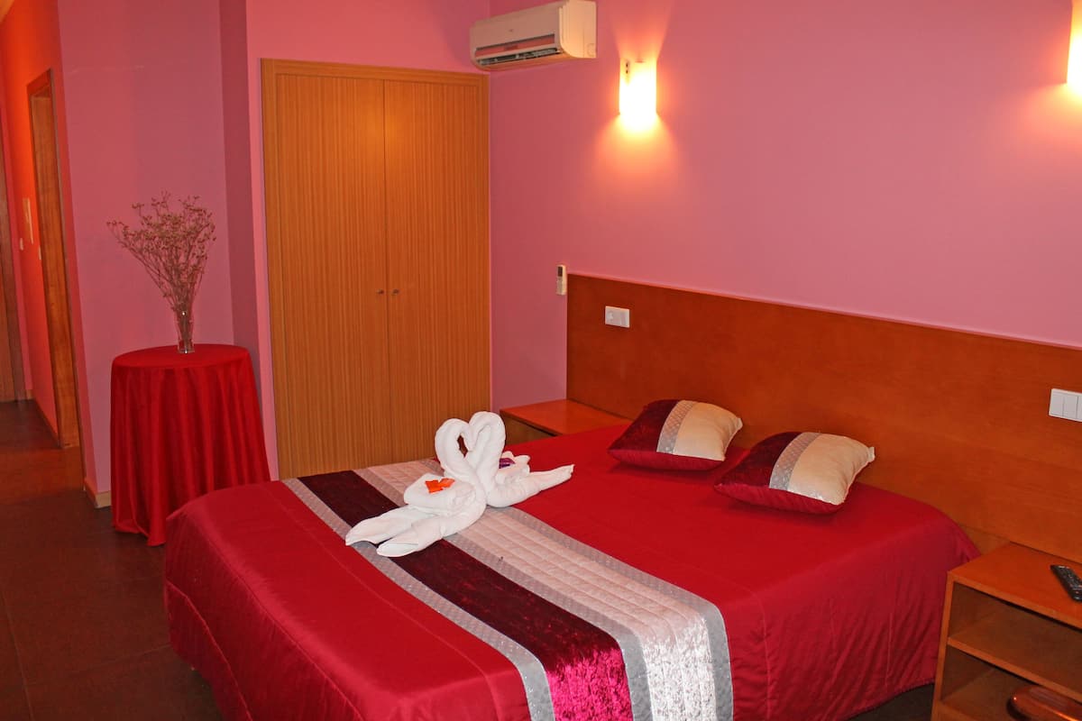 A hotel room with a red bedspread, a wooden headboard, a bedside table with a remote control, and a vase with flowers on a small table.