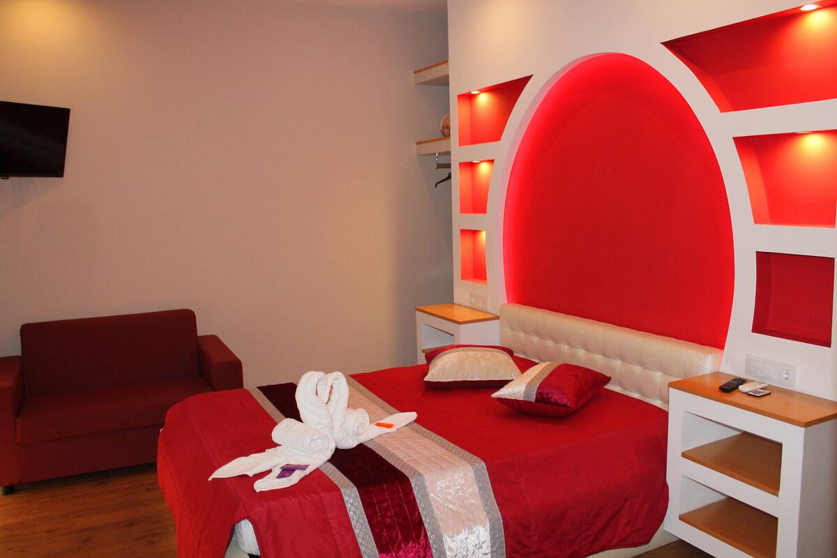 A hotel room with a red bed, a red sofa, a white nightstand, and a wall-mounted TV.