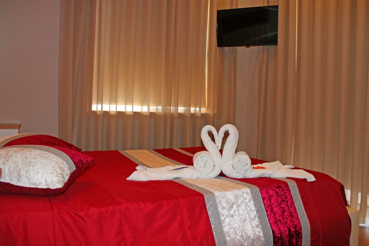 A hotel room with a red bedspread, two white pillows, and a pair of towels arranged in a heart shape on the bed.