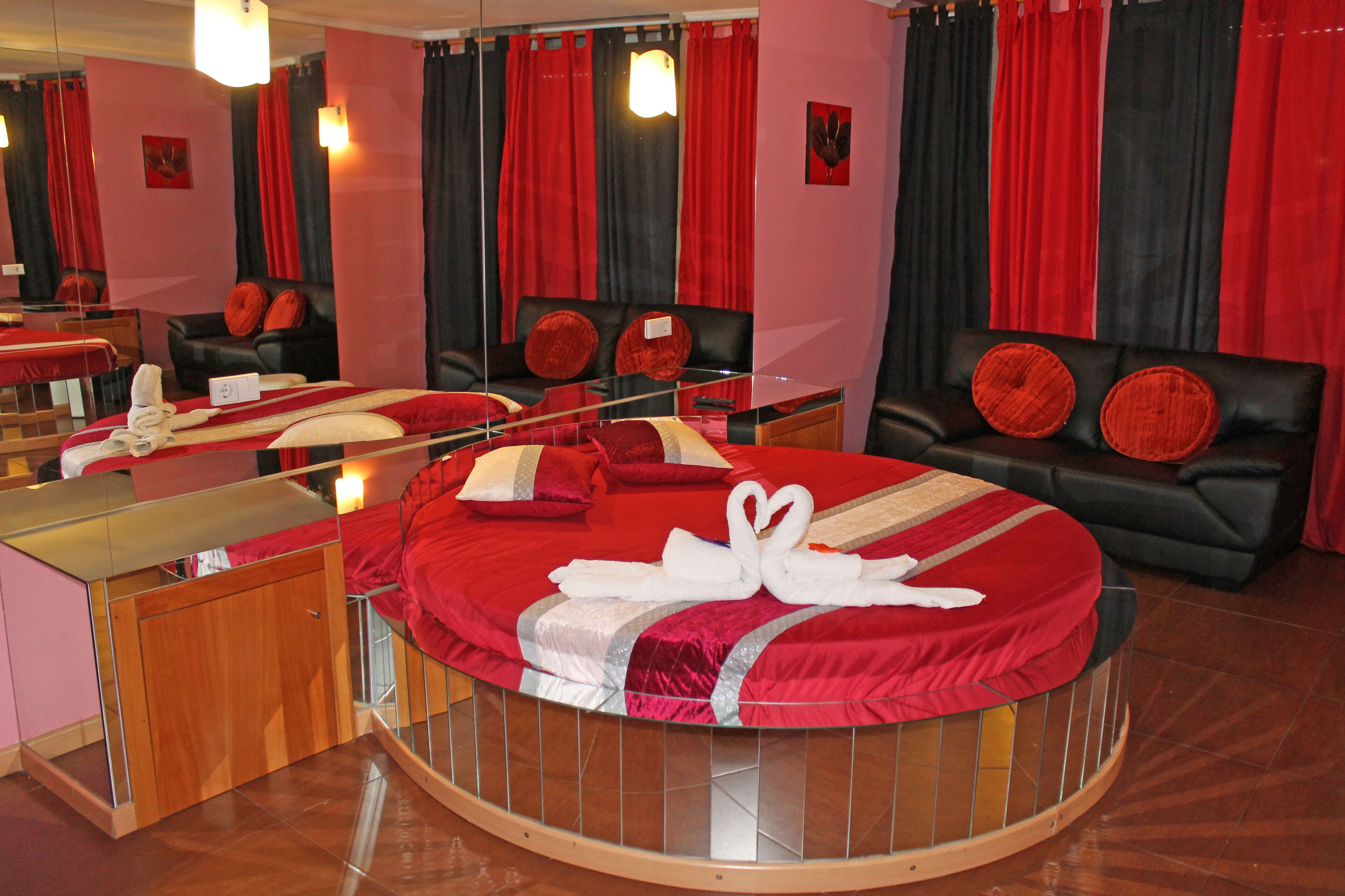A circular bed with red bedding and a mirror above it. There is a black leather sofa with red cushions in the background.
