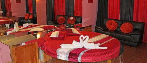 A circular bed with red bedding and a mirror above it. There is a black leather sofa with red cushions in the background.