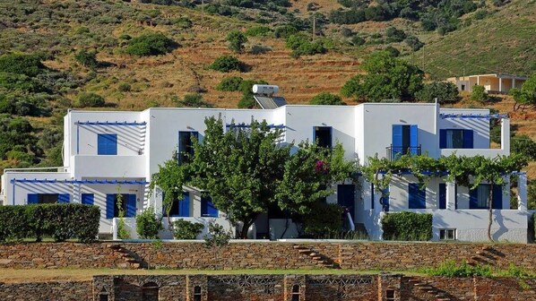 A white building with blue shutters, a tree in front, and a stone wall in the foreground. - Spanos Apartments (Andros)
