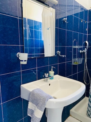 A bathroom with blue tiles, a white sink, a mirror, a towel, and a shower. - Spanos Apartments (Andros)