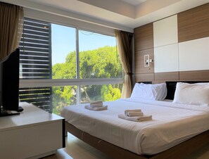 A modern hotel room with a large bed, a desk, and a view of trees through the window.