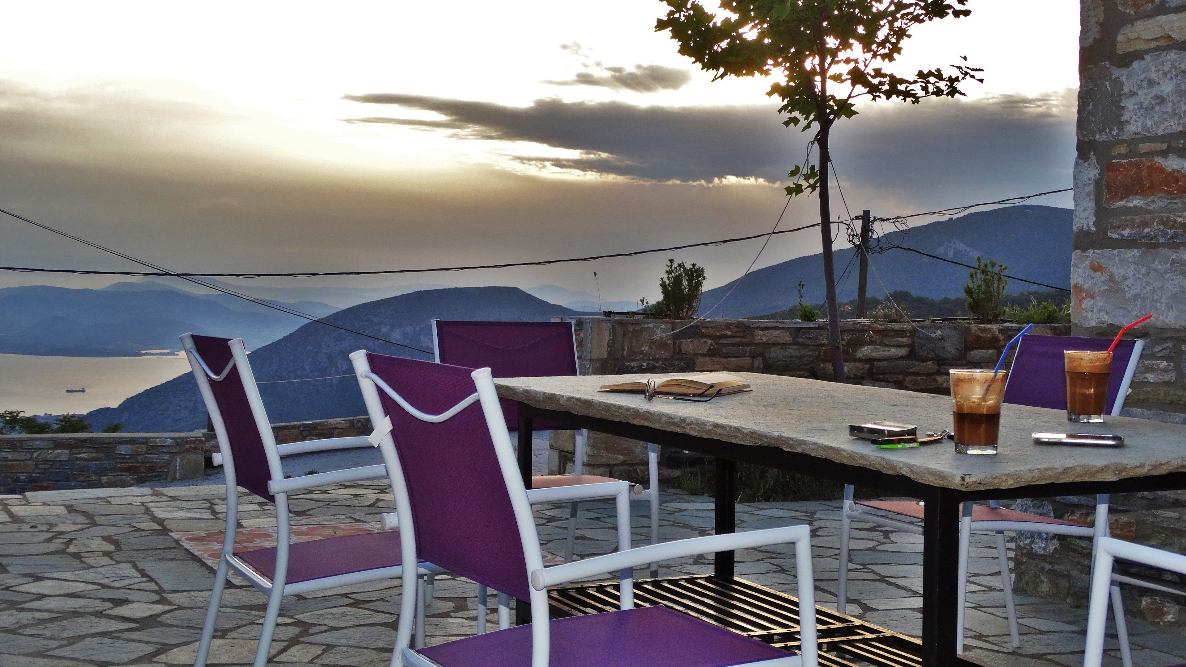 An outdoor table with chairs, a book, and drinks, set against a backdrop of mountains and a body of water.