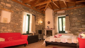 A cozy room with a stone wall, wooden beams, a fireplace, a bed, a red sofa, and a television. - Pelion Goddess Villas (Volos)