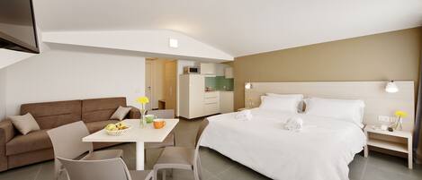 A modern hotel room with a bed, a sofa, a dining table, and a kitchenette.