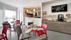 Cozinha moderna com TV, máquina de café e área de jantar com cadeiras vermelhas e mesa posta para dois.