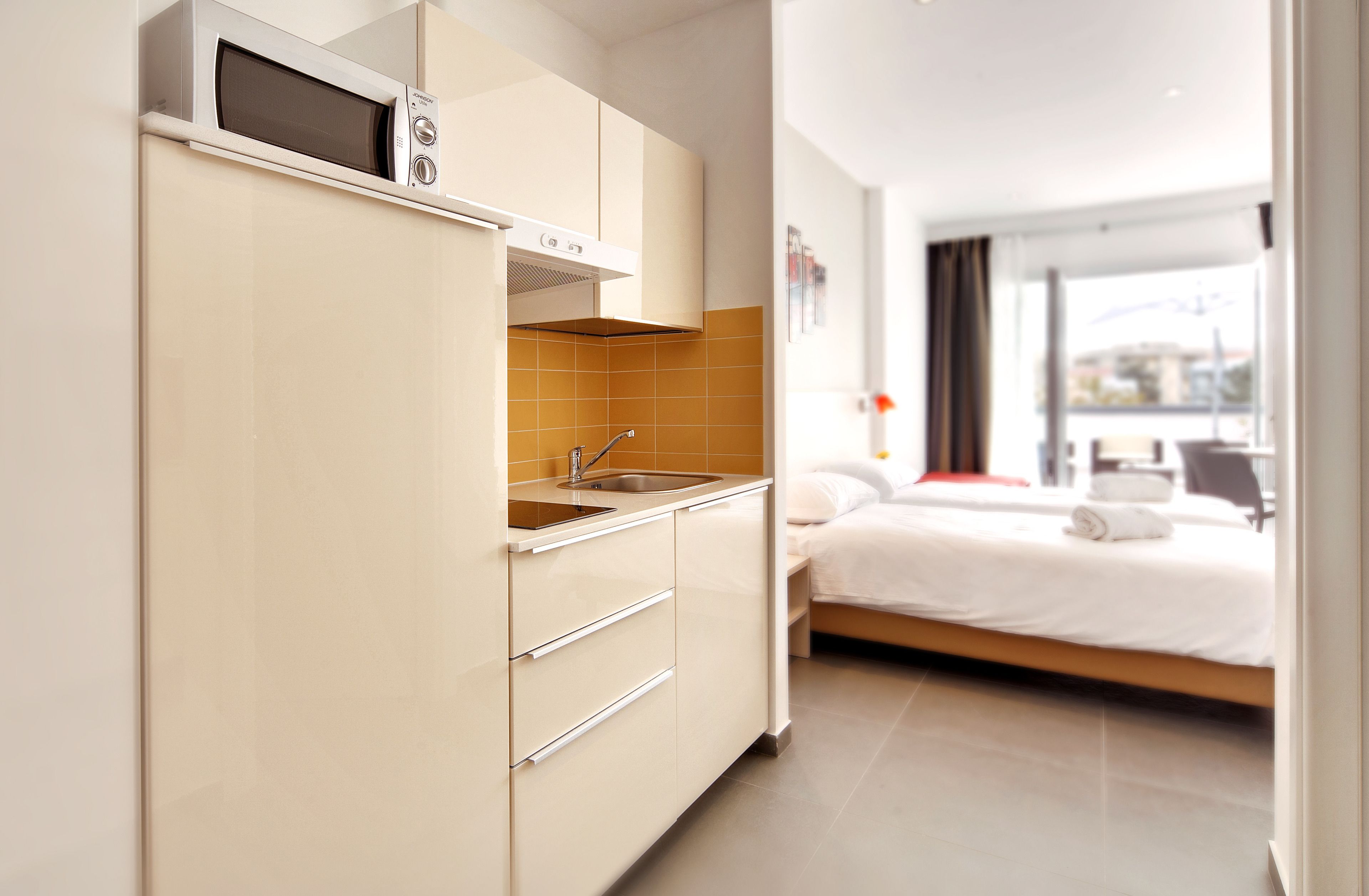A compact kitchen with a microwave, sink, and cabinets, adjacent to a bed with a view of the city.