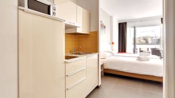 Een compacte keuken met een magnetron, spoelbak en kastjes, naast een bed met uitzicht op de stad.