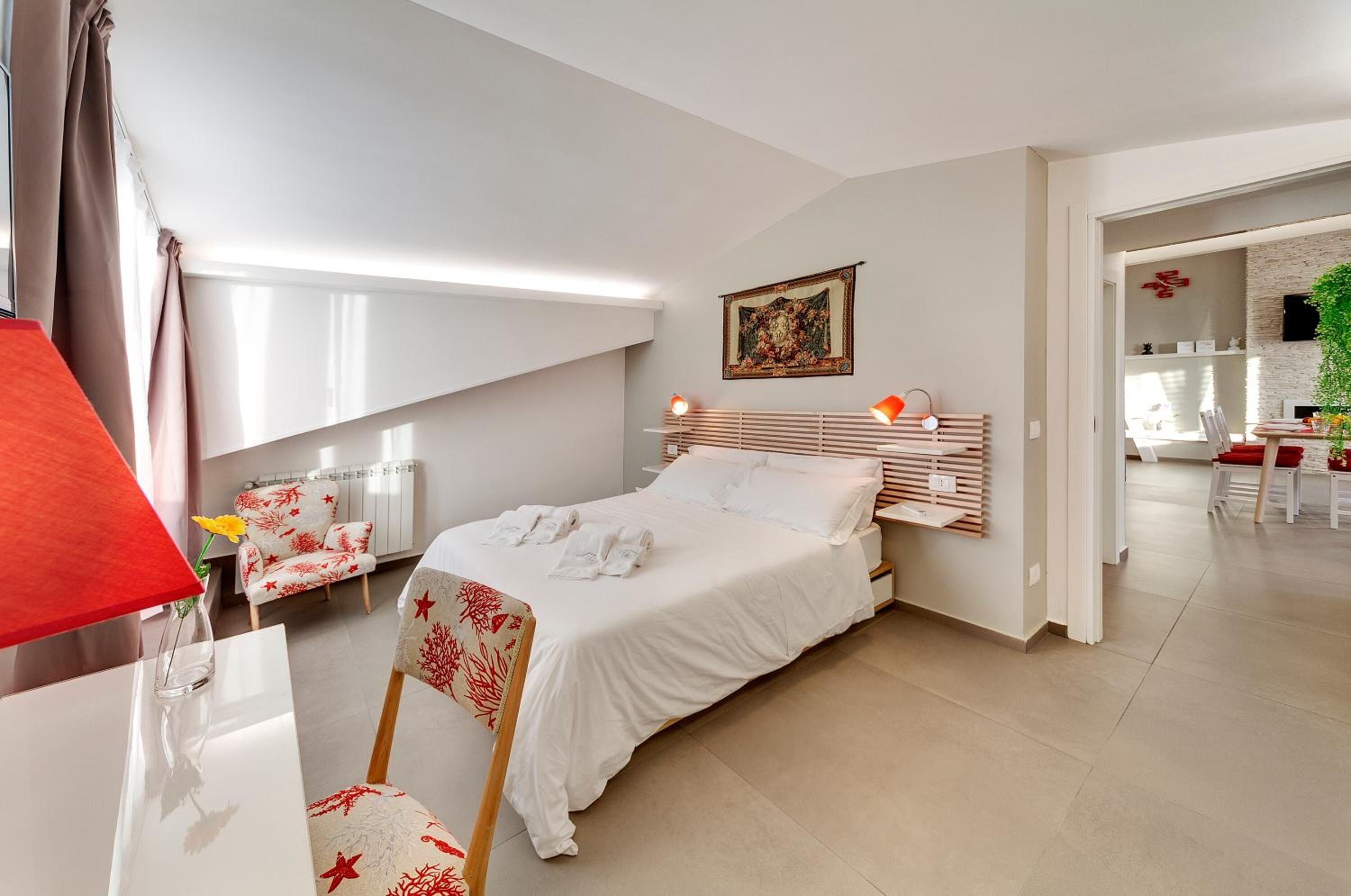 A modern hotel room with a large bed, a desk with a lamp, and a view of the dining area.
