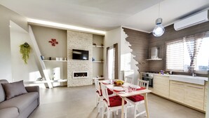 Uma sala de estar moderna com sofá, mesa de jantar e cadeiras. Há uma lareira, televisão e área de cozinha com armários e uma janela.