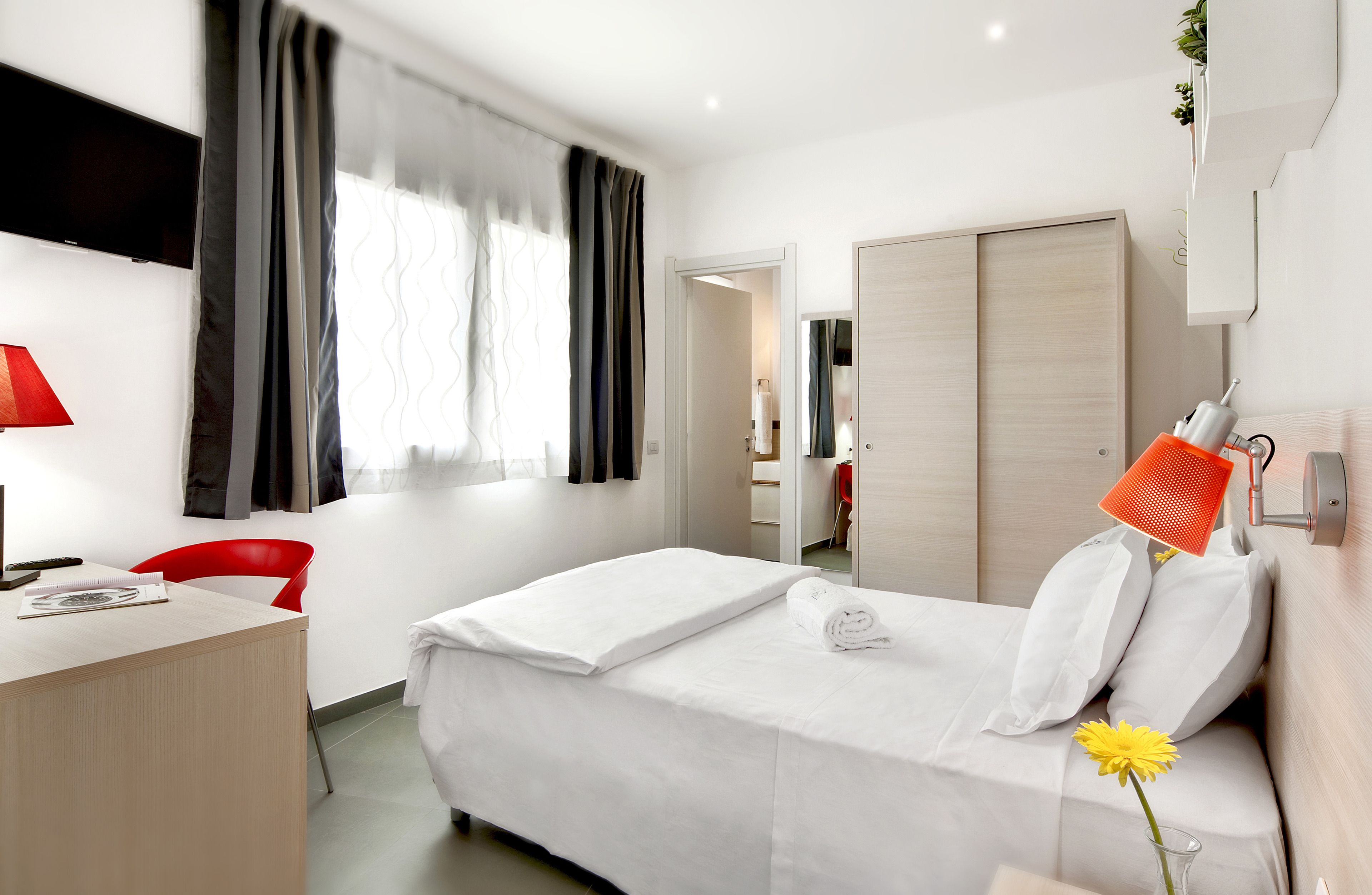 Habitación de hotel con cama, escritorio, silla, televisor y ventana con cortinas.