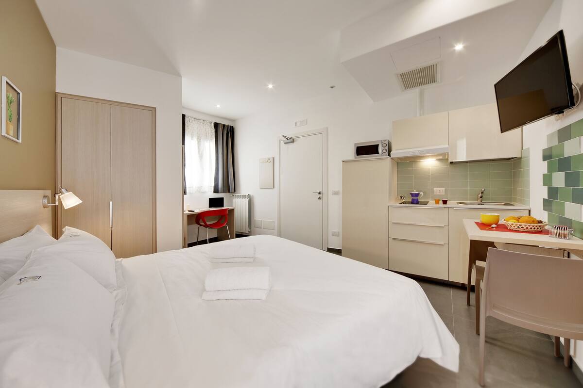 Ein Hotelzimmer mit einem großen Bett, einem Schreibtisch, einem Sessel, einem Fernseher und einer Küchenzeile.