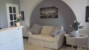 A living room with a sofa, two patterned cushions, a small table with a plant, a framed picture on the wall, and a white door. - B&B Il Cappero (Lipari)