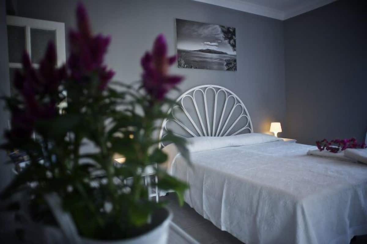 Un dormitorio con una cama blanca, una silla blanca, una mesita de noche con una lámpara y un cuadro colgado en la pared.